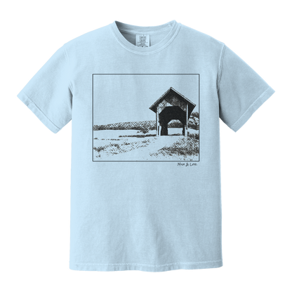 Front view of the Covered Bridge cotton t‑shirt in Chambray (baby blue), with a storybook scene of a rustic covered bridge arching over a quiet Vermont dirt road.