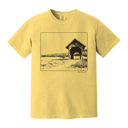 Front view of the Covered Bridge cotton t‑shirt in Mustard (yellow), with a storybook scene of a rustic covered bridge arching over a quiet Vermont dirt road.