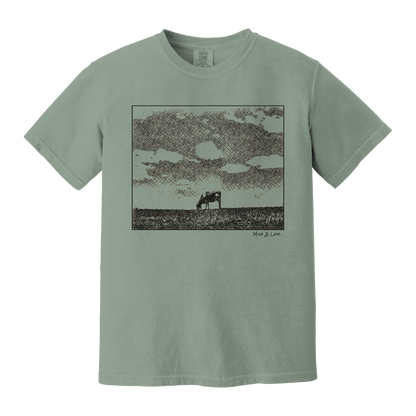 Front view of the Field Cow Cotton T-Shirt in Bay (green) featuring  a whimsical black-and-white rendering of a lone cow grazing in a field beneath a cloud-dotted sky