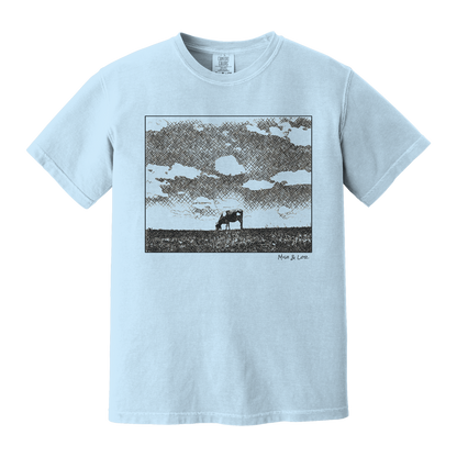 Front view of the Field Cow Cotton T-Shirt in Chambray (baby blue) featuring  a whimsical black-and-white rendering of a lone cow grazing in a field beneath a cloud-dotted sky
