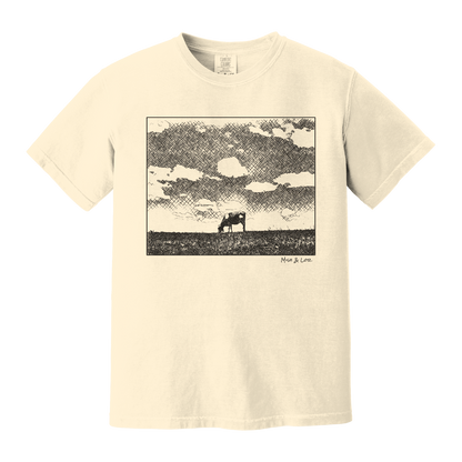 Front view of the Field Cow Cotton T-Shirt in Ivory (cream) featuring  a whimsical black-and-white rendering of a lone cow grazing in a field beneath a cloud-dotted sky